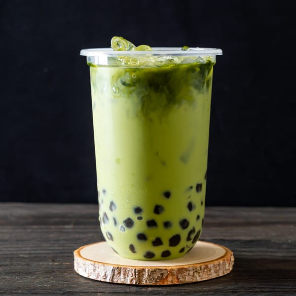 Green matcha boba tea with ice in a clear plastic cup on a wooden coaster.