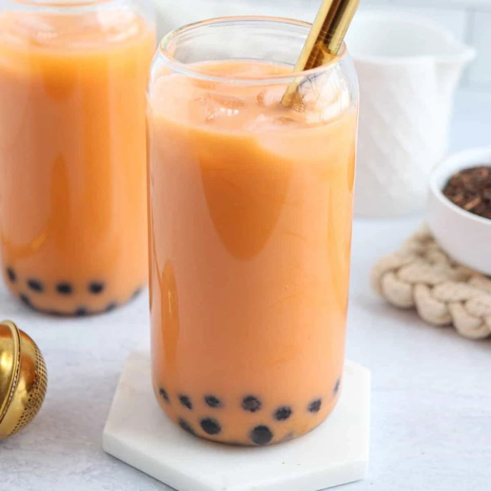 Two glasses of orange Thai iced tea with boba pearls and a gold straw.
