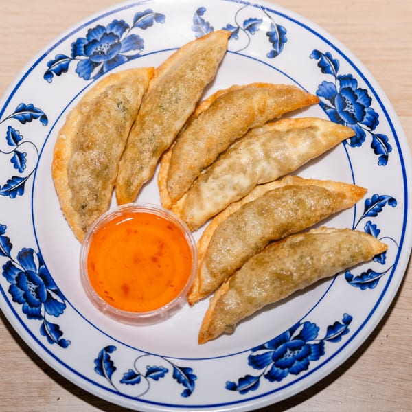 Seven golden-brown fried dumplings with a side of orange dipping sauce on a white and blue floral plate.